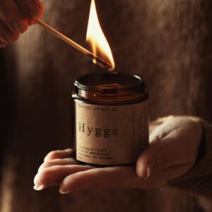A hand lights a Hygge candle, creating a warm and cozy atmosphere indoors.