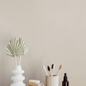 White creative vase with decorative artificial plants and organic soap with body brush placed near set of bamboo toothbrushes in ceramic holder and dark glass reusable bottle of lotion
